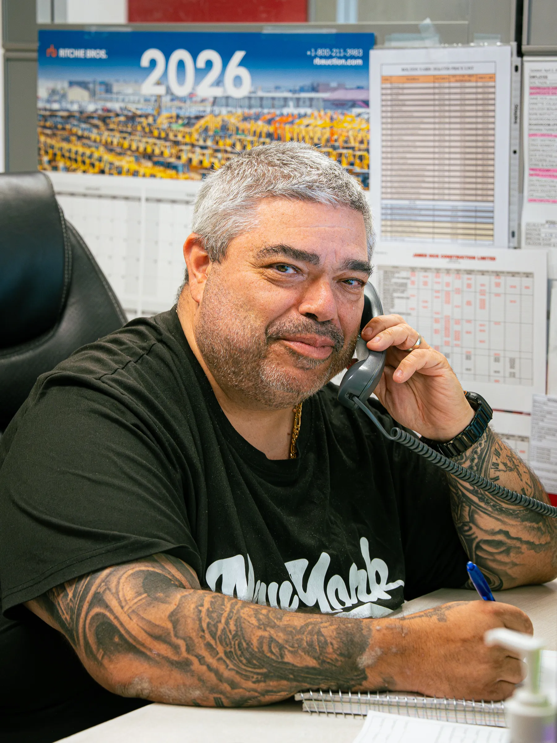 JDA team member on a desk phone in the dispatch office, with scheduling charts and a calendar on the wall behind him