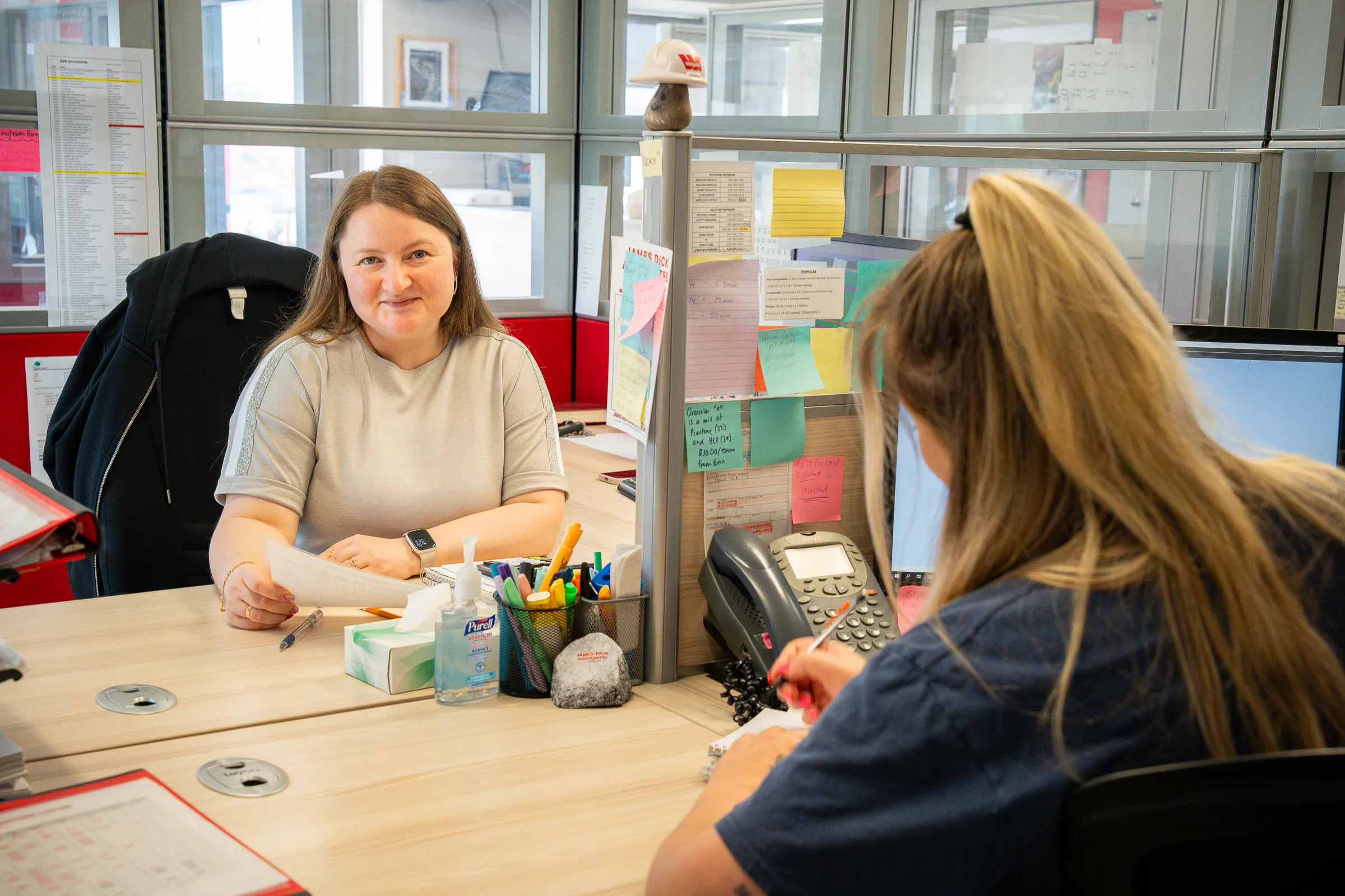 Two JDA office team members talking across their shared desk in a bright windowed workspace